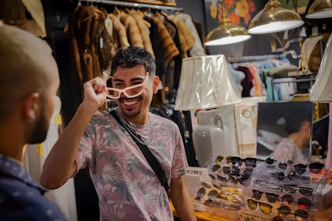 Two people in a vintage clothing shop. A man holds up sunglasses while smiling. Racks of clothing, lamps, and sunglasses displays surround them.
