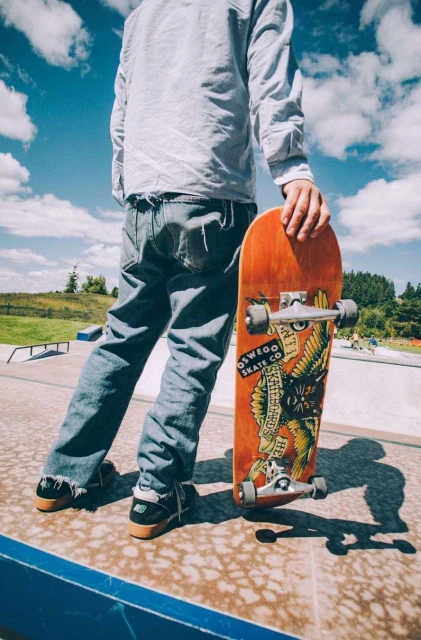 Person stands at a skatepark, holding a skateboard with graphic art, wearing jeans, against blue sky and cloud background.