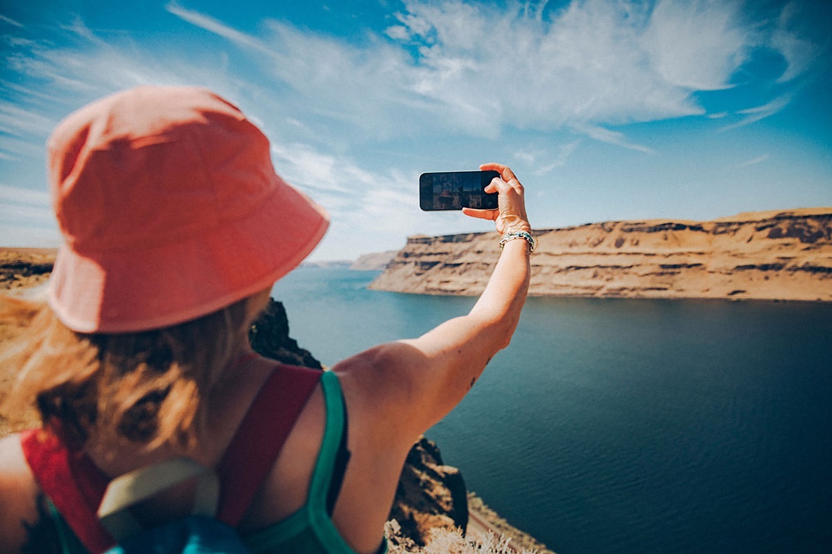Adult female takes a smartphone photo of a canyon lake landscape, wearing a hat and backpack on a sunny day with blue sky and white clouds.