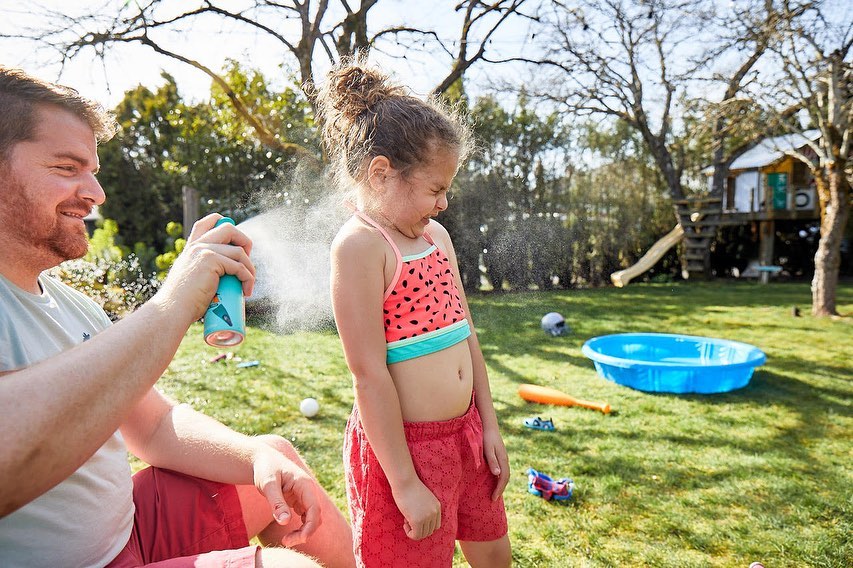 Father applying sunscreen to a young girl in a watermelon swimsuit in a sunny backyard with grass, trees, and a playset.