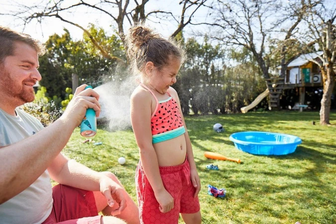 Father applying sunscreen to a young girl in a watermelon swimsuit in a sunny backyard with grass, trees, and a playset.