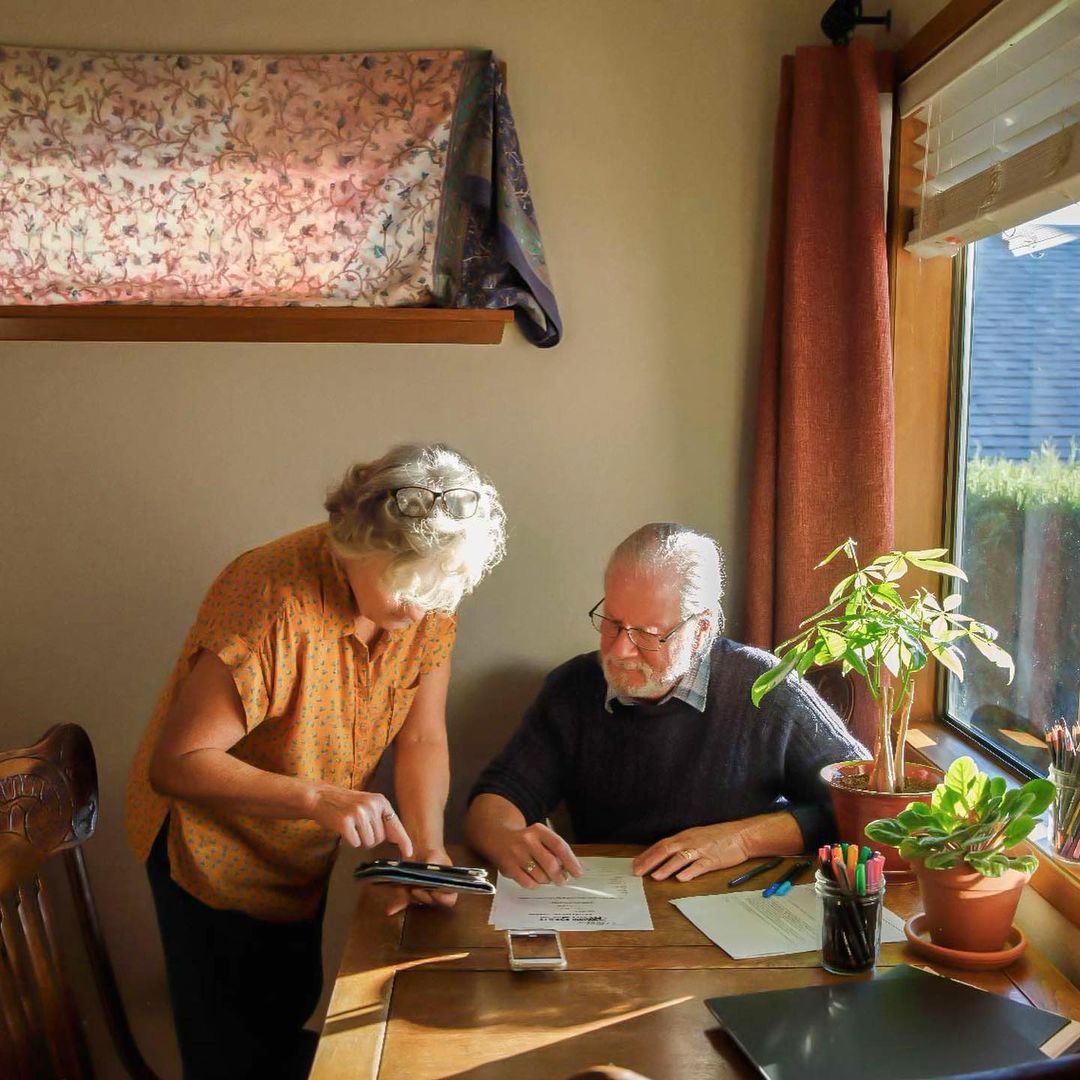 Elderly couple working at a wooden table indoors. Woman in orange shirt uses phone while man points to paper in natural light.