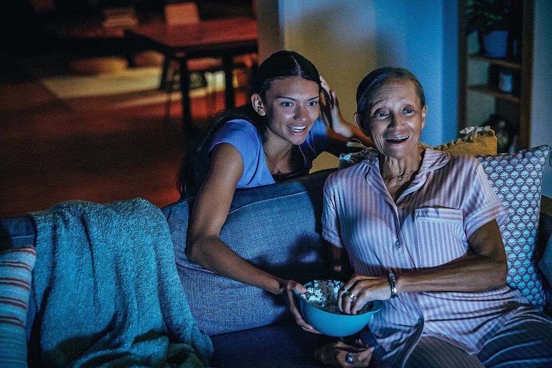 An adult woman and senior adult woman sit on a couch together eating popcorn in an indoor living room setting with soft lighting.