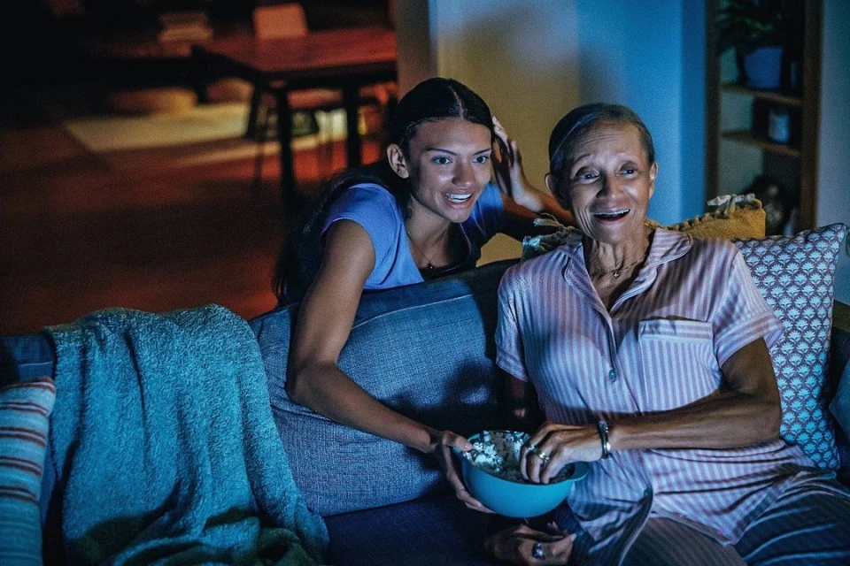 An adult woman and senior adult woman sit on a couch together eating popcorn in an indoor living room setting with soft lighting.