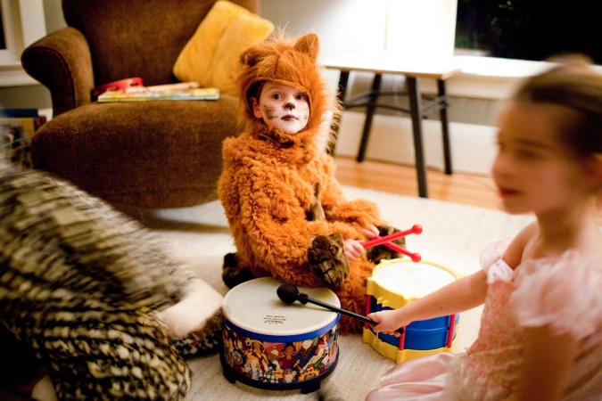 A young child in a furry animal costume plays a drum with red drumsticks, alongside another child in a pink dress indoors.