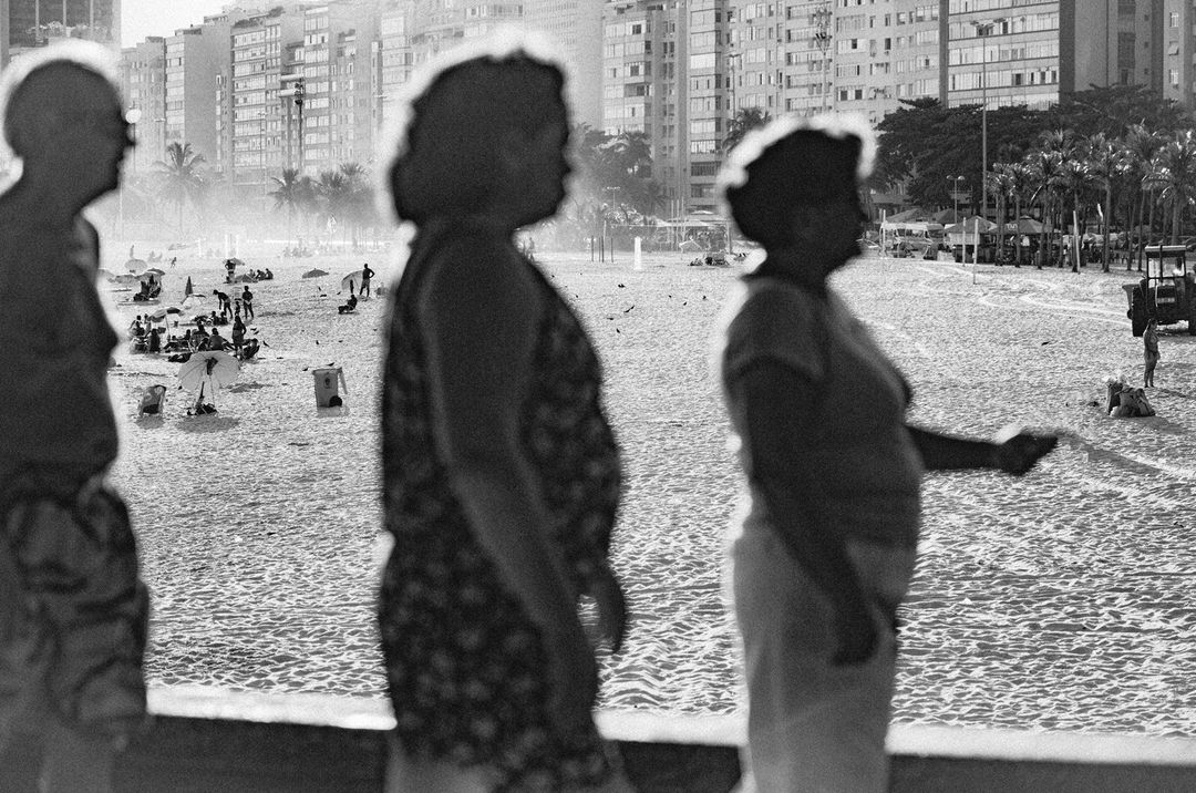 Black and white shot of three people standing on a sandy beach with apartment buildings and palm trees in the background.