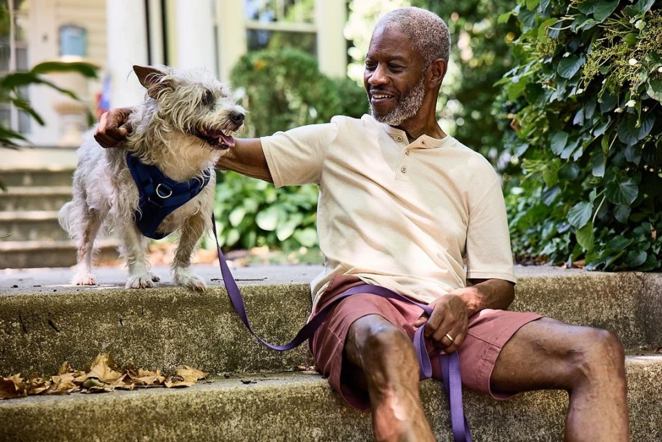 Smiling African-American man sits with dog on steps outdoors. Dog wears harness, man has dog leash; trees and house in background.