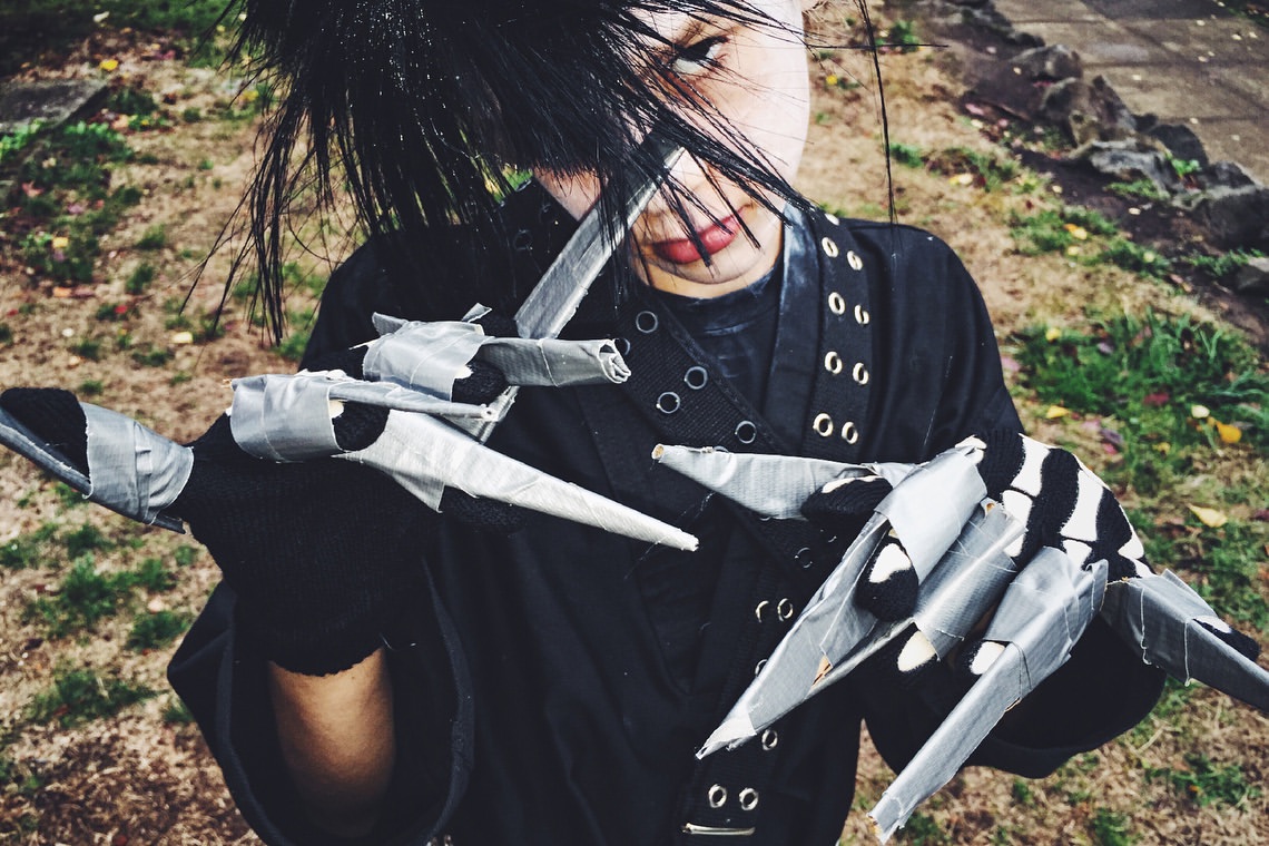 Person in a creative Edward Scissorhands costume with razor finger blades, black clothing, and wild black hair, outdoors with foliage.