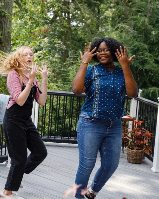 Two women posing outdoors in casual clothing on a deck surrounded by greenery, smiling and gesturing with their hands raised.
