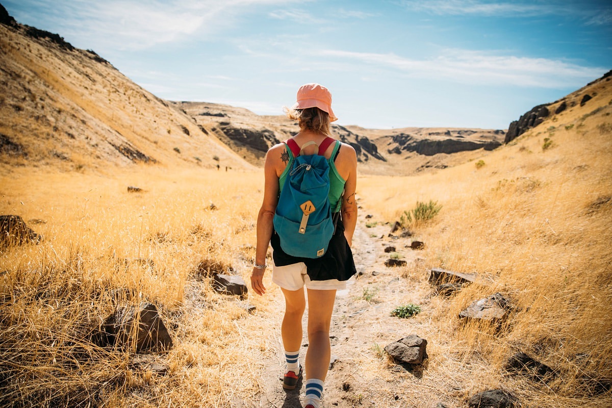 Woman hikes a desert trail wearing a bucket hat, backpack, and athletic wear under a blue sky with tall, dry grass and canyon walls.