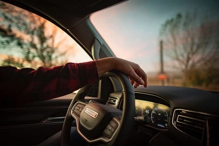 Interior view of a car dashboard and steering wheel with driver's hand, showcasing sunset through the windshield, creating a warm, inviting atmosphere.