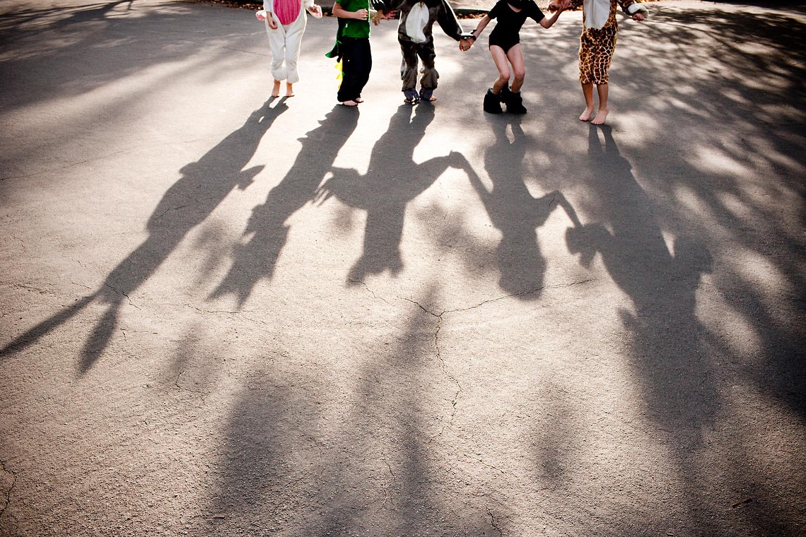 Five children in animal costumes hold hands, casting long shadows on a grey concrete surface in an outdoor, sunny setting. 