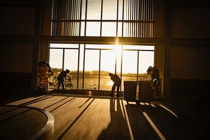 Airport scene with silhouettes of people near a large window, bright sunlight streaming through, casting long shadows across the floor.
