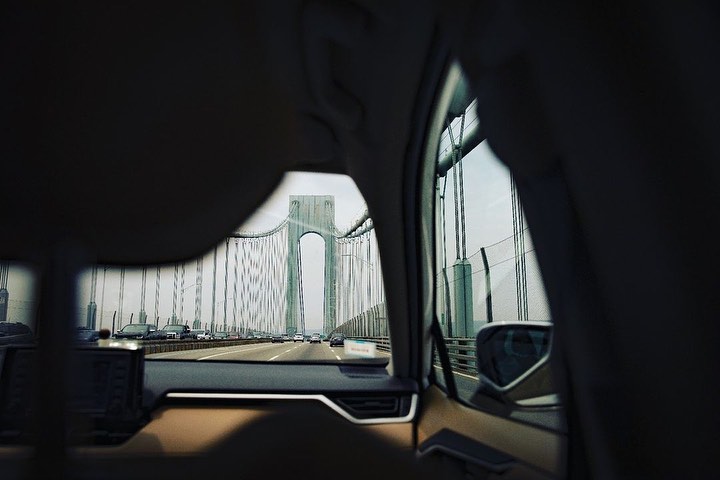 View of the Verrazano-Narrows Bridge from inside a vehicle on a highway, capturing the bridge's structure and surrounding cars in daylight.