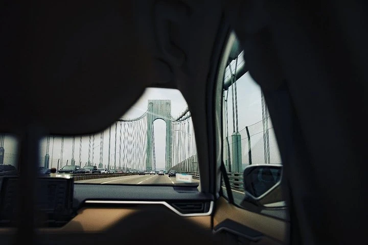 View of the Verrazano-Narrows Bridge from inside a vehicle on a highway, capturing the bridge's structure and surrounding cars in daylight.