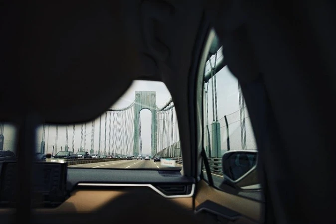 View of the Verrazano-Narrows Bridge from inside a vehicle on a highway, capturing the bridge's structure and surrounding cars in daylight.