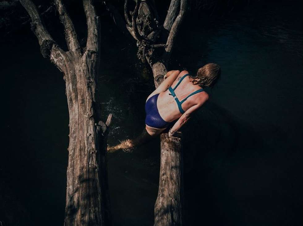 Back view of a woman sitting on a tree trunk, wearing a blue swimsuit, with a body of water in the background.