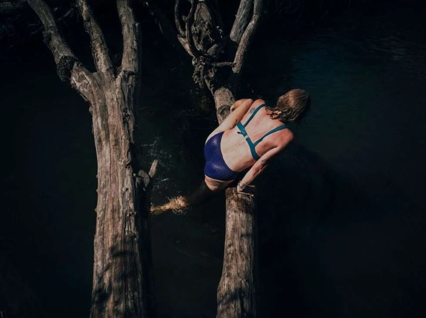Back view of a woman sitting on a tree trunk, wearing a blue swimsuit, with a body of water in the background.