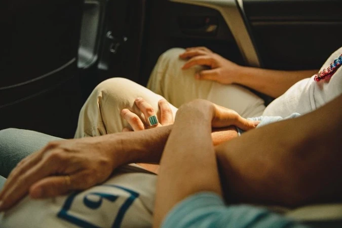Close-up shot of two people sitting in a car, with hands resting on lap. One person has a ring with a large green stone.