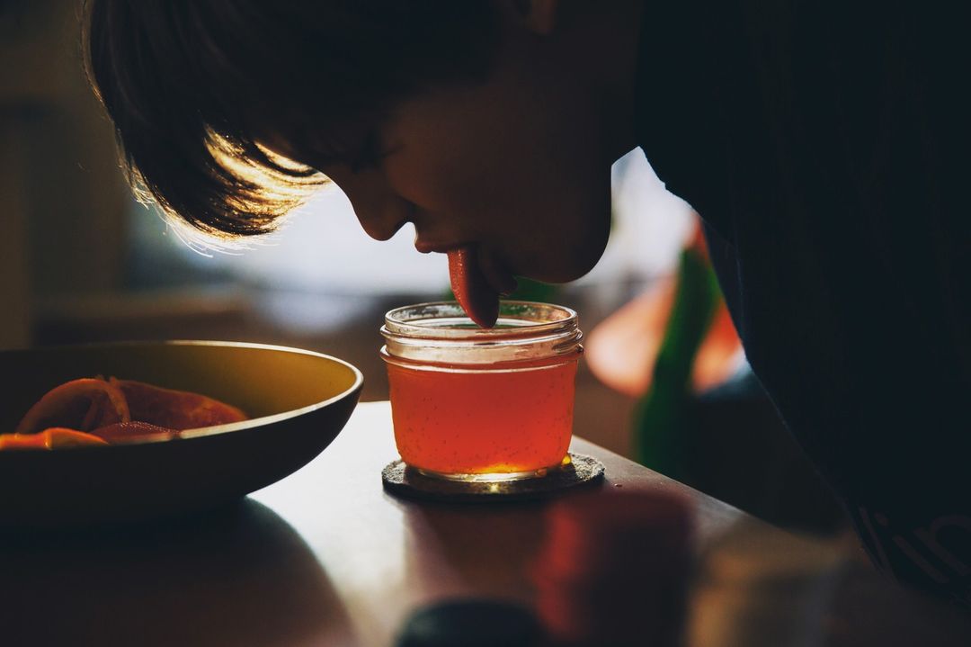 A close shot features a person drinking red liquid from a mason jar, with a bowl of fruit alongside on a wood surface indoors.