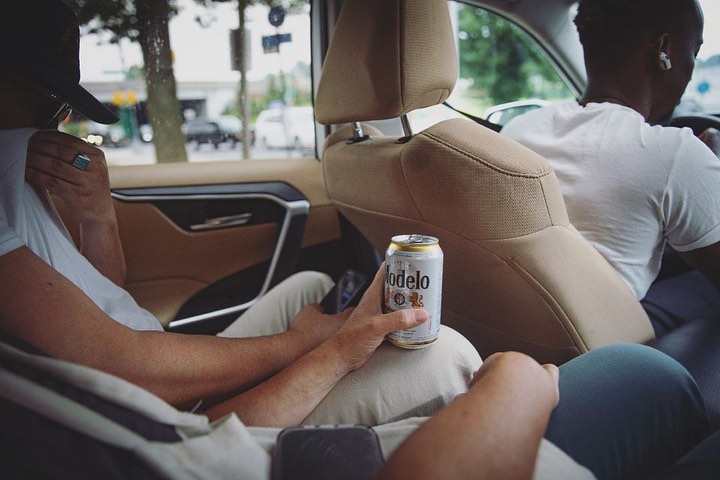 Two adult men are passengers in a car. One holds a Modelo beer can, while the other is driving. The interior of the vehicle is tan.
