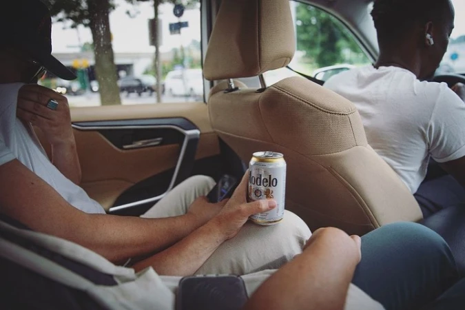 Two adult men are passengers in a car. One holds a Modelo beer can, while the other is driving. The interior of the vehicle is tan.
