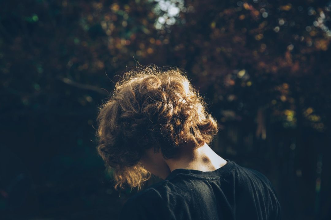 Back of a person's head with curly red hair wearing a dark shirt, in a shadowy outdoor setting with blurred foliage.