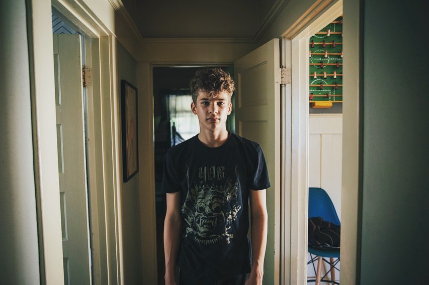 A young man with curly hair stands in a hallway, wearing a t-shirt with a graphic print, facing the camera in an indoor setting.
