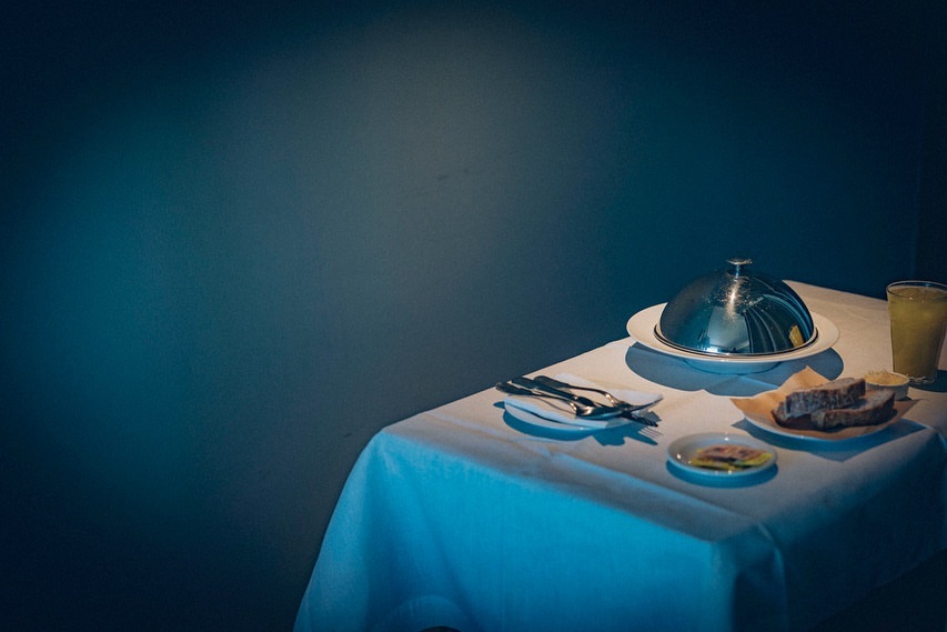 Elegant table setting with domed serving dish, bread, and drink, set against a dark teal backdrop, creating a contrast in light and color.