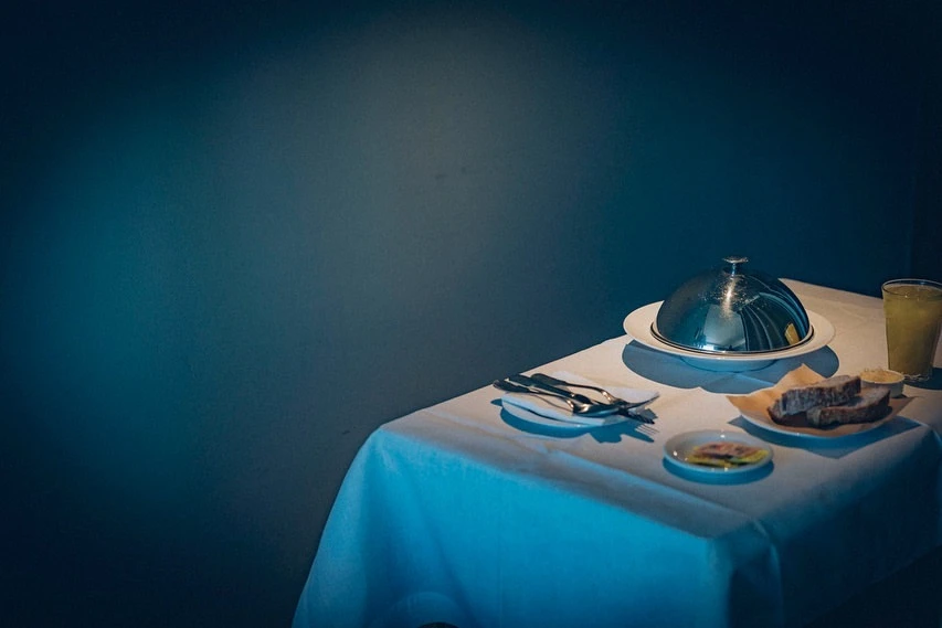 Elegant table setting with domed serving dish, bread, and drink, set against a dark teal backdrop, creating a contrast in light and color.