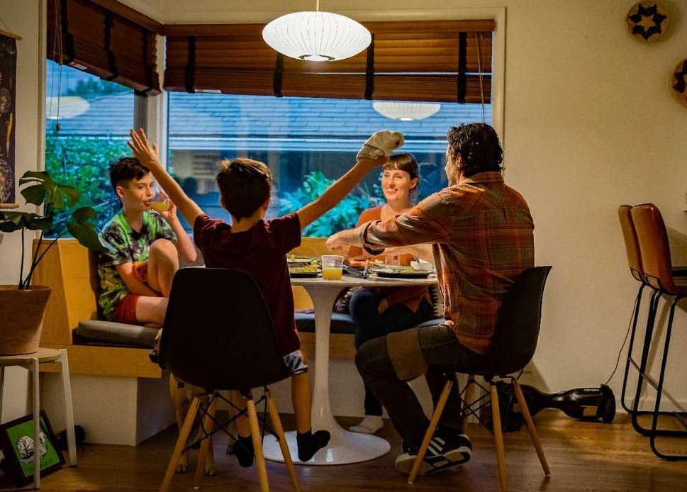 Family of four enjoys a meal together at a round white table in their home dining area, with light from a pendant lamp overhead.