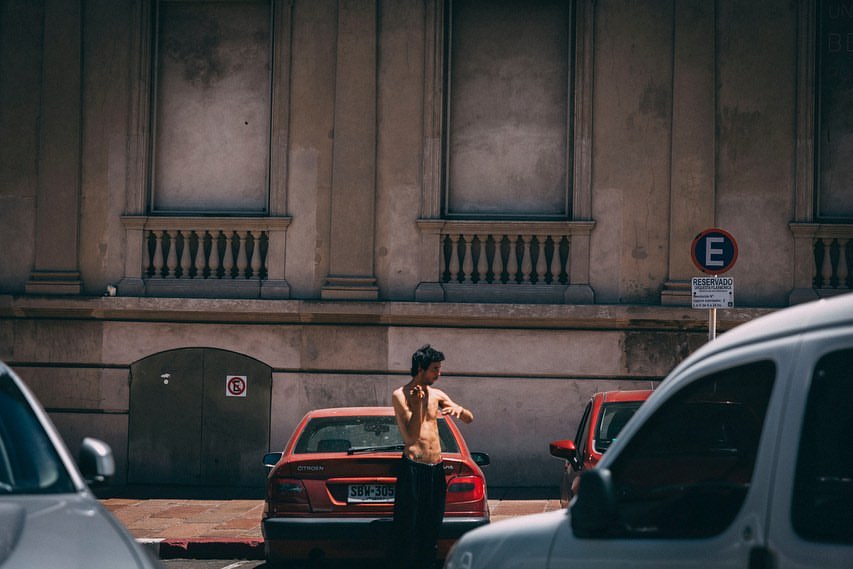 Young man standing shirtless by a red car in an urban environment on a sunny day with a building in the background and other cars around.