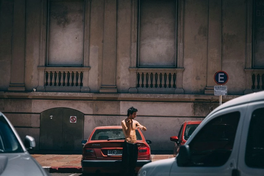 Young man standing shirtless by a red car in an urban environment on a sunny day with a building in the background and other cars around.