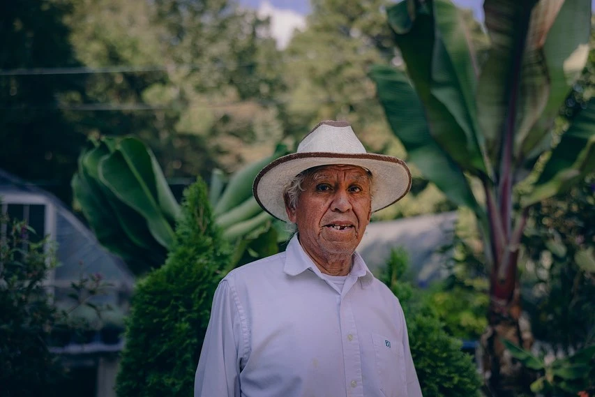 Elderly man in a white shirt and hat poses outdoors surrounded by lush greenery, captured in a medium shot with natural lighting.