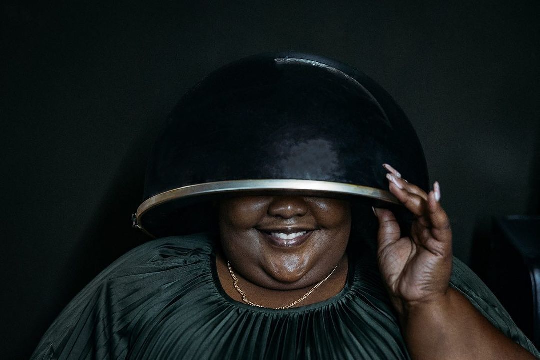 Adult female smiles with black hair dryer helmet on her head in a beauty salon, wearing a green dress in a studio shot.