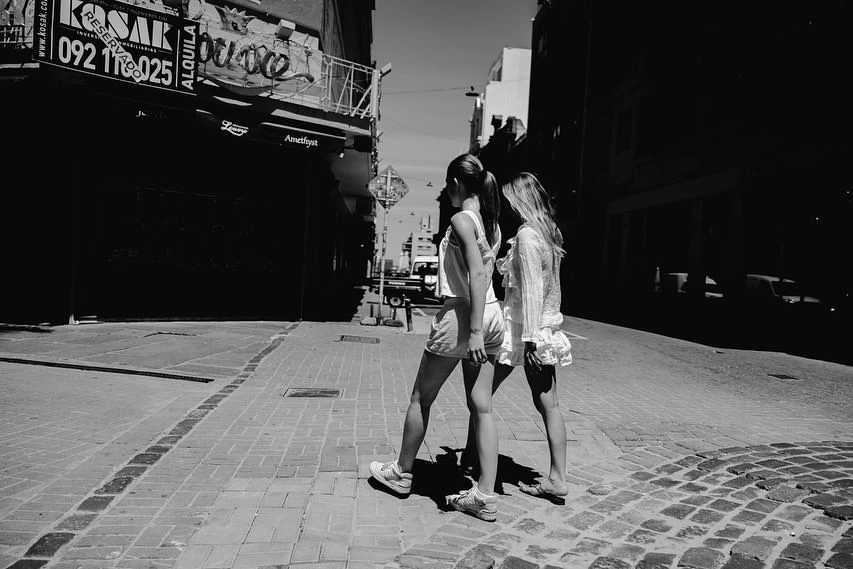 Two young women in summer outfits walk on a cobblestone street on a sunny day, captured in black and white with shadows.