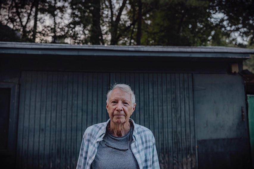 Portrait of an elderly man with gray hair wearing a plaid shirt, standing in front of a gray building and trees.