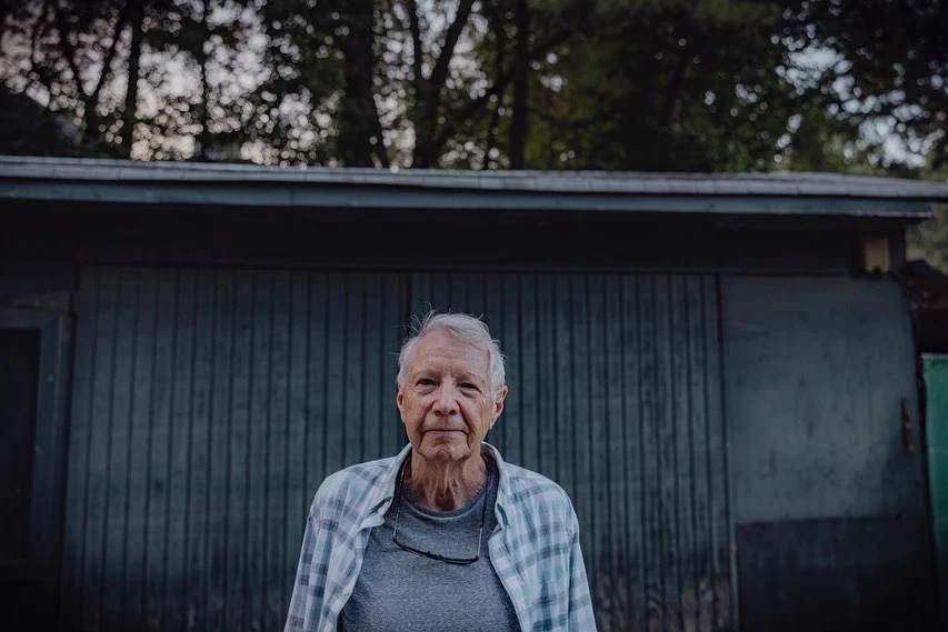 Portrait of an elderly man with gray hair wearing a plaid shirt, standing in front of a gray building and trees.
