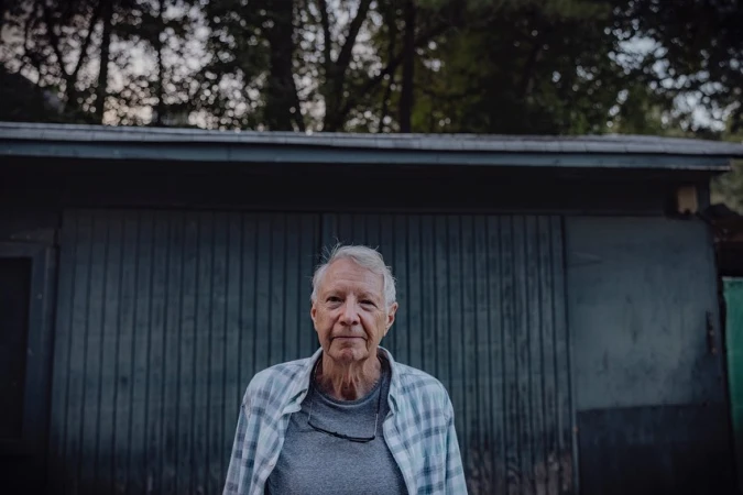 Portrait of an elderly man with gray hair wearing a plaid shirt, standing in front of a gray building and trees.