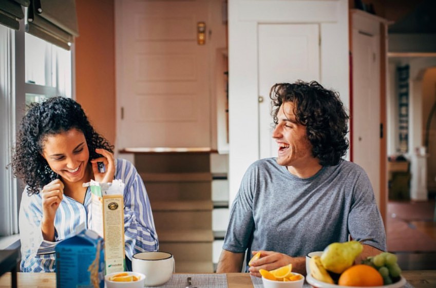 Two people laugh at a table with fruit and juice. The woman wears pajamas and the man a t-shirt in a brightly lit home setting.