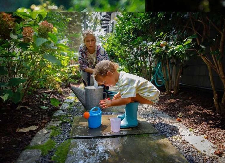 A child plays with water in a garden with a woman supervising, featuring plants, watering cans, and a stone pathway in natural light.