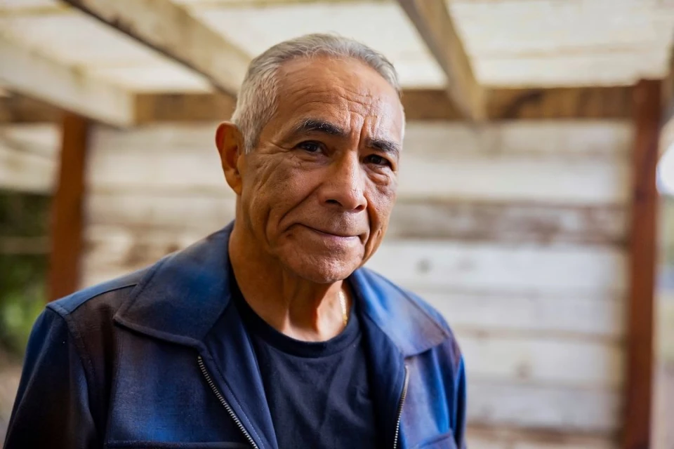 Portrait of an elderly man with short gray hair in a navy jacket smiling indoors against a blurred light-brown wooden background.