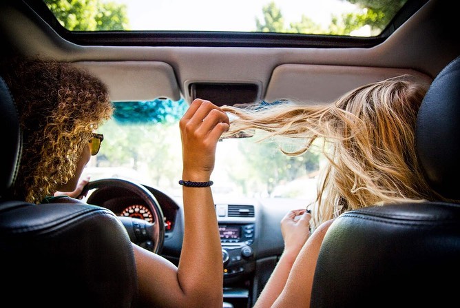 Two women in a car with an open sunroof, one driving while the other plays with her hair, showcasing a light and airy aesthetic.