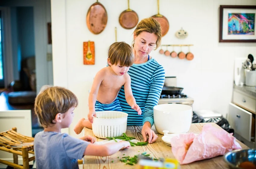 A woman and two children are in a kitchen, preparing food, with copper pots hanging on the wall and natural light coming through.