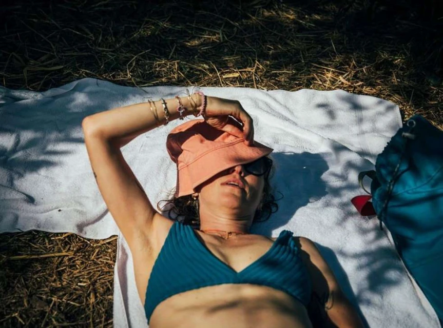 Woman in teal bikini top lies on a white towel in the sun, wearing sunglasses and a peach bucket hat partially covering her eyes.