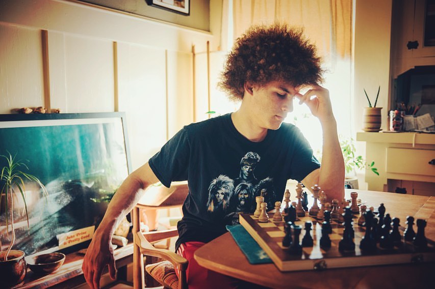 Young man with curly hair sits indoors, pondering a chess board on a wooden table, wearing a band t-shirt in diffused lighting.