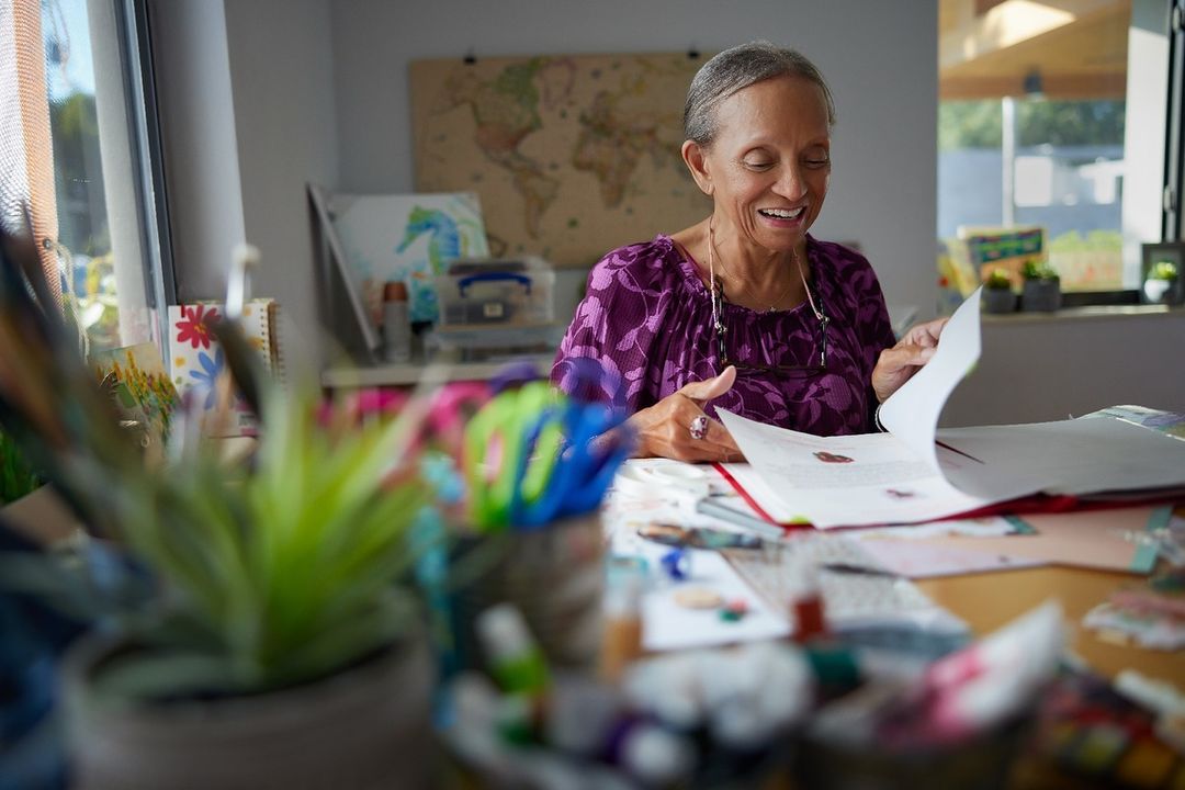 Smiling mature woman seated at table in a brightly lit room, reviewing documents amongst colorful art supplies and drawing materials.