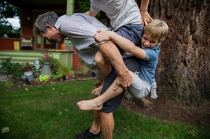 Man carrying a child on his back in a backyard with a house and tree in the background