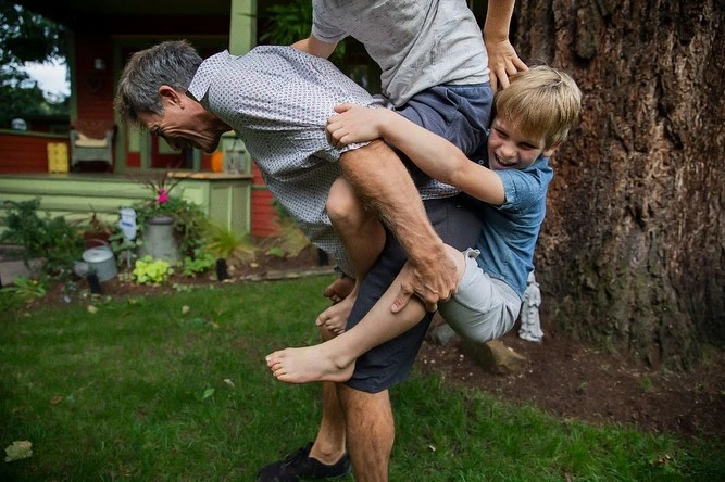 Man carrying a child on his back in a backyard with a house and tree in the background