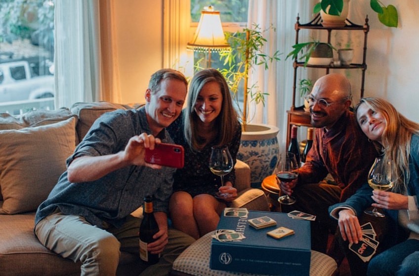 Four adults smiling indoors while a man takes a photo with his phone, with a board game and wine bottles on the table in front of them.